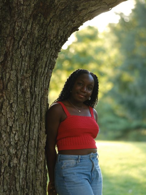 Image of incoming MSU Honors College student and Williams Scholar Kiran Ofei-Amoh softly smiling and leaning against a large tree, wearing a vibrant red top and blue jeans. Blurred greenery is in the background.