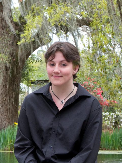 Image of incoming MSU Honors College student and Williams Scholar Jordan Penfold softly smiling in front of a pond, large tree, and flowers. Penfold is wearing a black collared shirt.