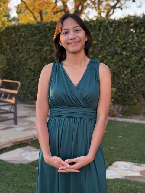 Image of incoming MSU Honors College student and Williams Scholar Milee Zarate-Bayani standing and softly smiling in a long teal dress. Trimmed hedges and a bench are in the background.
