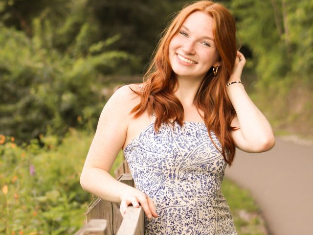Honors Research Scholar Devin Olep smiling and posing in a sundress. Lush greenery is in the background.