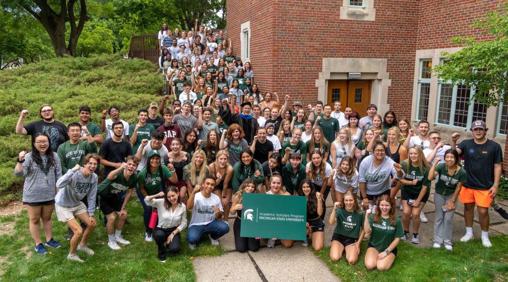 **Alternative text:** A large group of approximately 120 incoming Michigan State University Academic Scholars Program students gathers outside the MSU Alumni Chapel for the program's annual Welcome & Convocation. Students, many dressed in green and white Spartan apparel, fill the chapel steps and surrounding walkway, smiling, cheering, and raising their fists in celebration. Several students kneel in the front holding an Academic Scholars Program sign, while faculty and staff stand among the group. The photo captures the excitement, energy, and sense of community as the newest cohort begins their MSU journey.