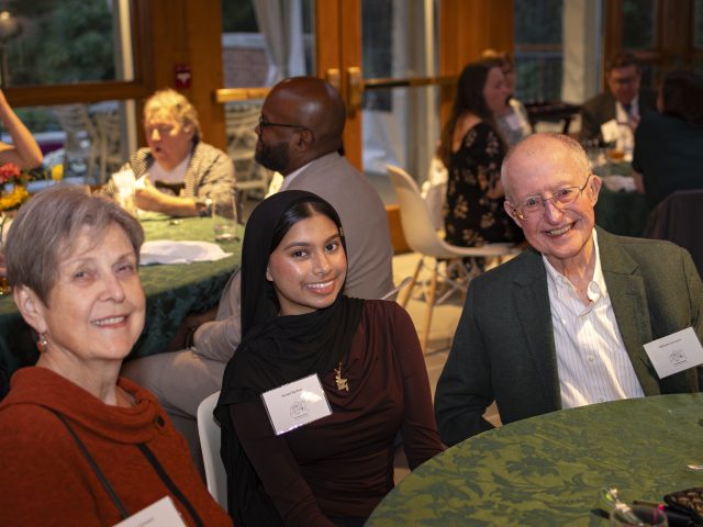 Sanaa Bashar sitting with Linda and Bill Schoenl at the Honors College Scholarship Dinner at a table. Behind them, others sit at other tables discussing with people around them.