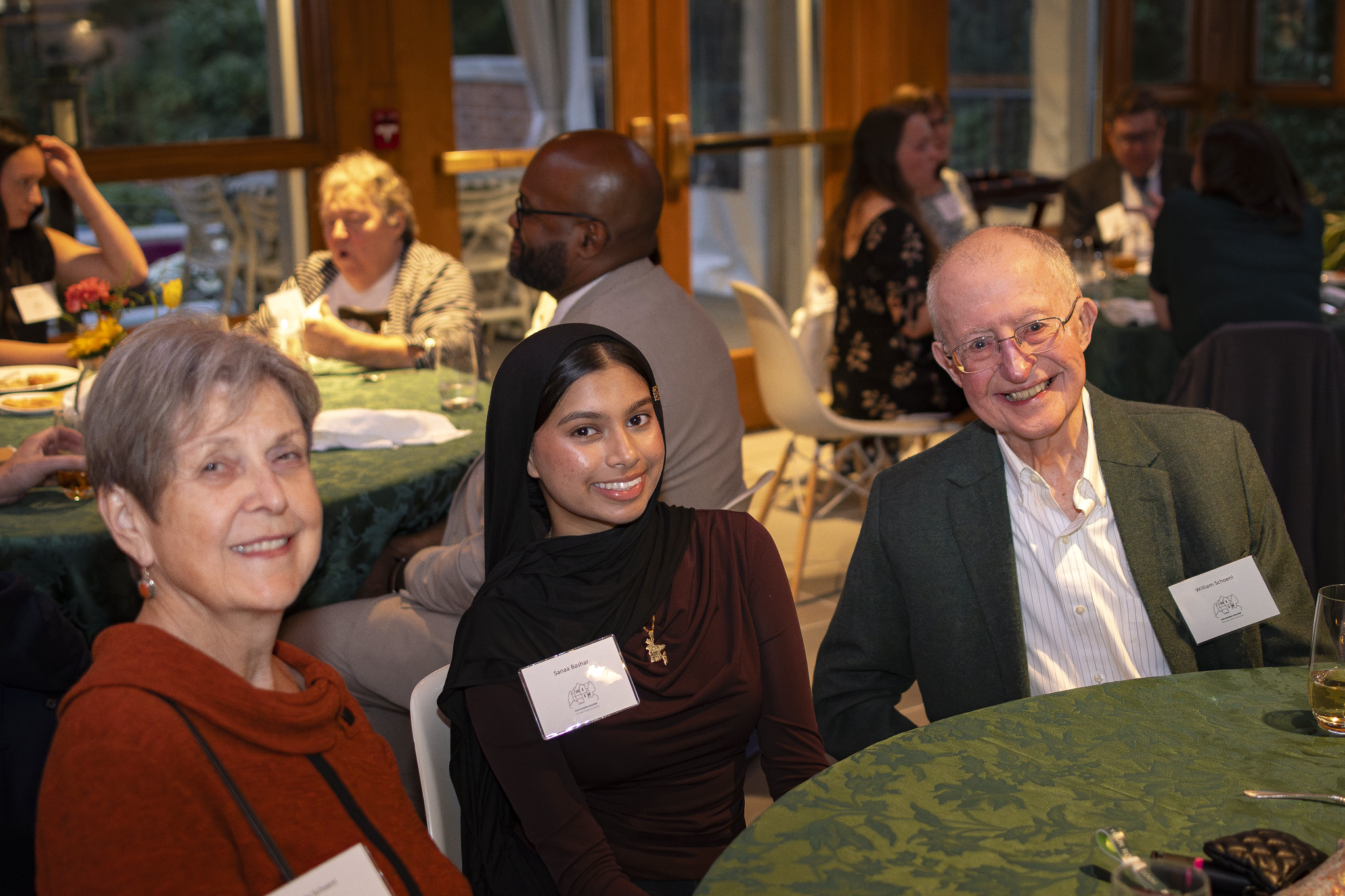 Sanaa Bashar sitting with Linda and Bill Schoenl at the Honors College Scholarship Dinner at a table. Behind them, others sit at other tables discussing with people around them.