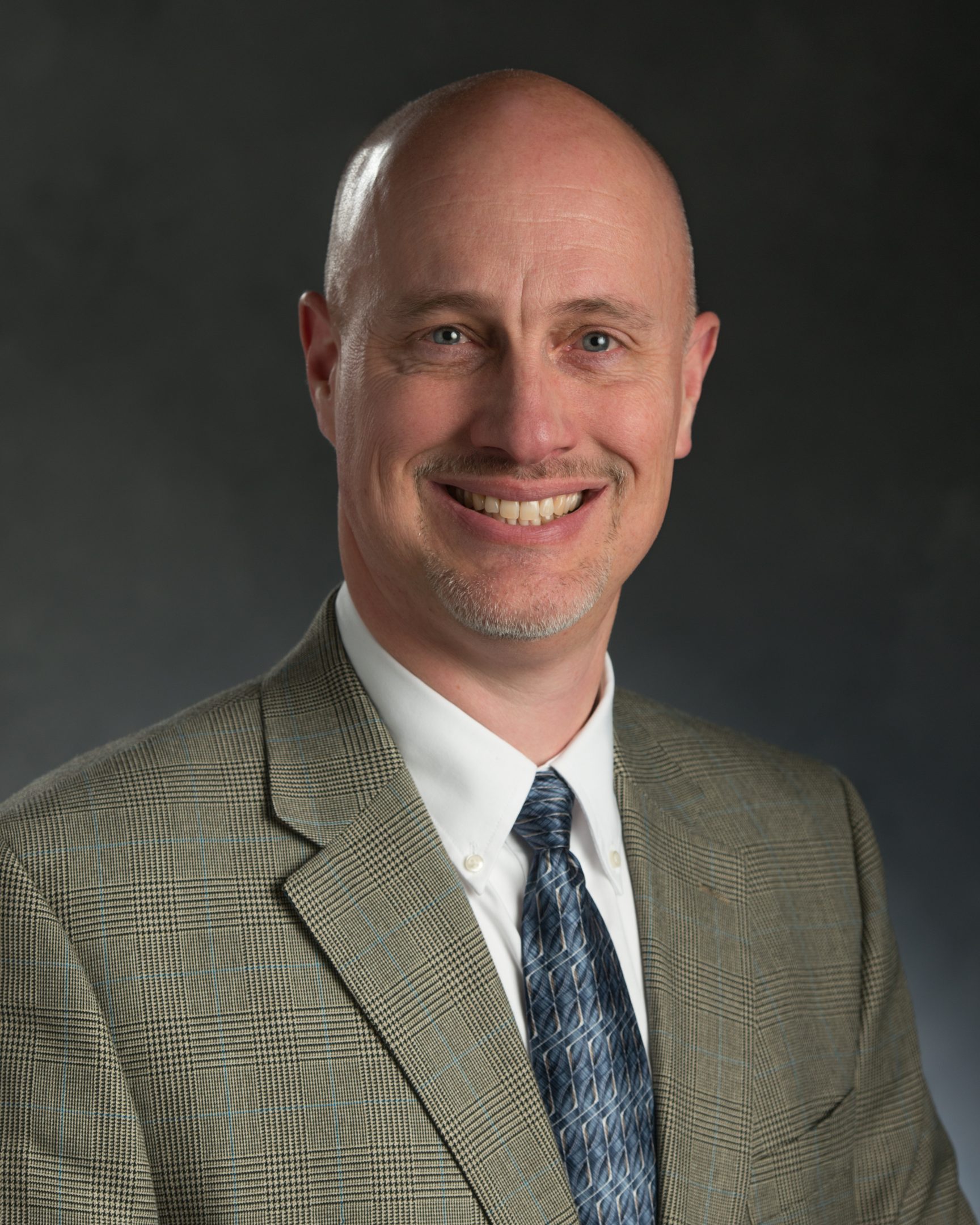 MSU Honors College alumnus Bradley Marks smiling in a dark tan suit jacket, white collared shirt, and blue patterned tie.