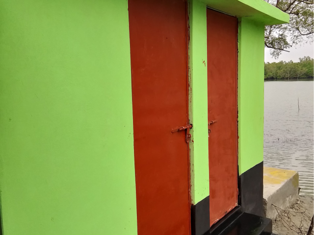 Completed latrine in Datinakhali, painted bright green with two reddish-brown doors and black trim, located beside a body of water.
