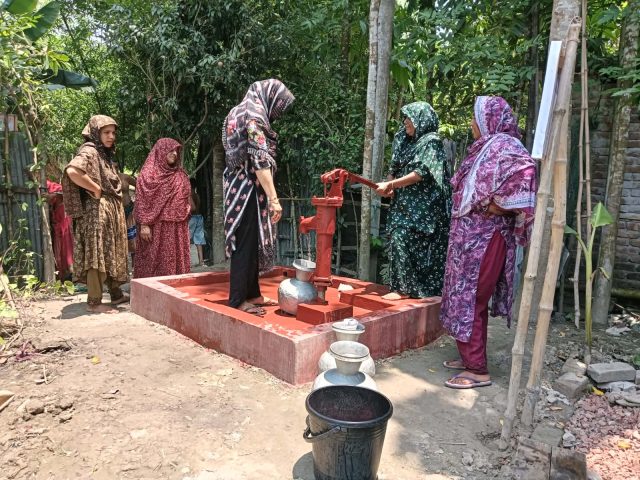 A group of residents surround a bright red tube well in Gobindapur, using it to pour water. Trees surround them.