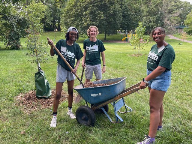 Three MSU Honors College students in HC IMPACT t-shirts standing and smiling with a wheelbarrow full of woodchips.