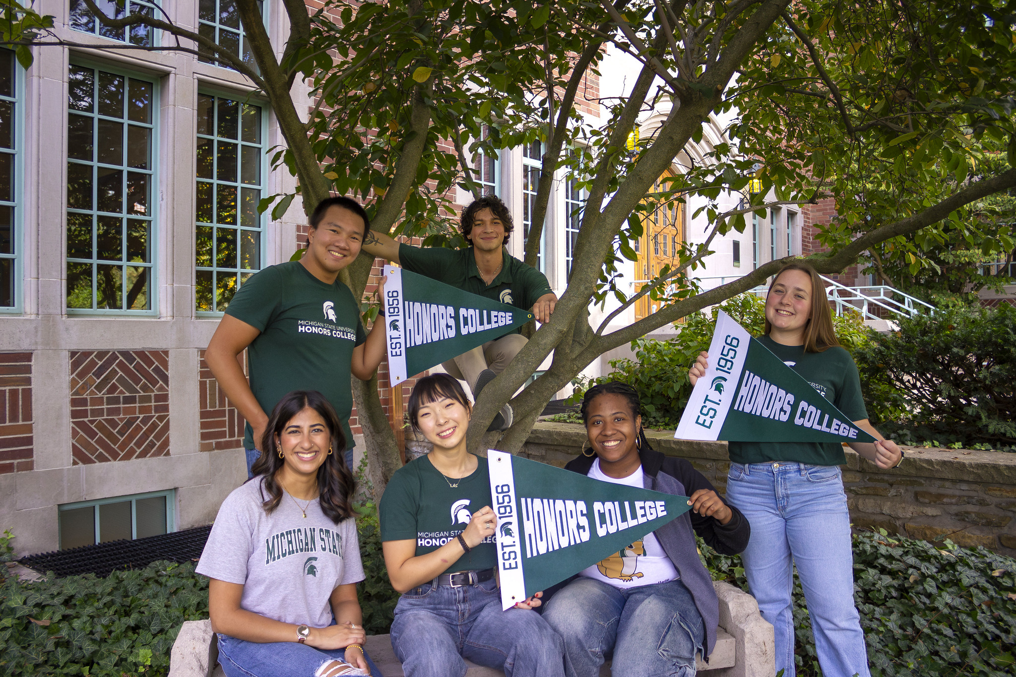 Six Honors College students in various Michigan State attire, smiling and posing outdoors with green and white Honors College pennants. Three of the students are sitting on a stone bench, two are standing, and one in crouched in a tree. Campbell Hall is in the background.
