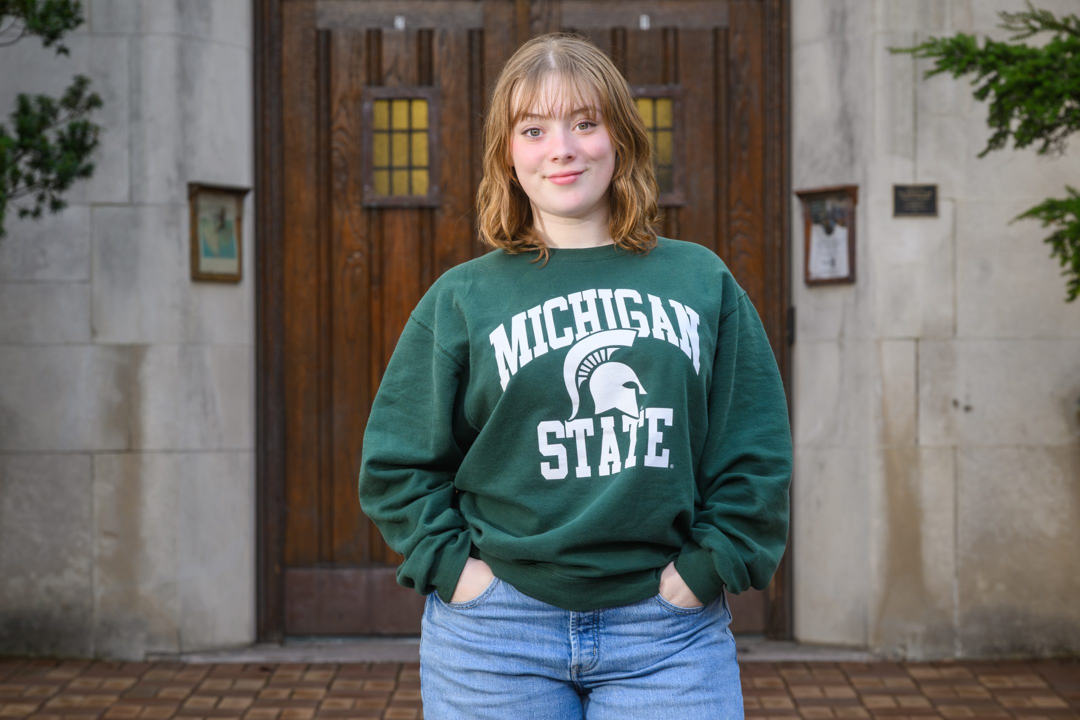 Honors College student Anastasia Breen standing and smiling in front of Beaumont Tower with hands in pockets, wearing a green and white Michigan State sweater.