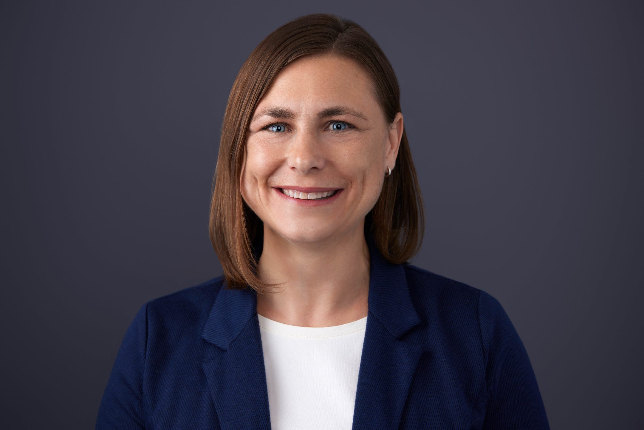 Michigan State University Honors College alumna Dru Montri smiling in a white top and navy blazer against a dark grey gradient background.