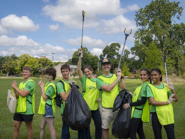 A group of seven MSU Honors College students in neon vests and green t-shirts posing in their trash pickup gear for HC IMPACT.