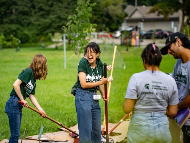 An MSU Honors College student laughing joyfully while holding a gardening tool and doing community service with a group of other HC IMPACT participants.
