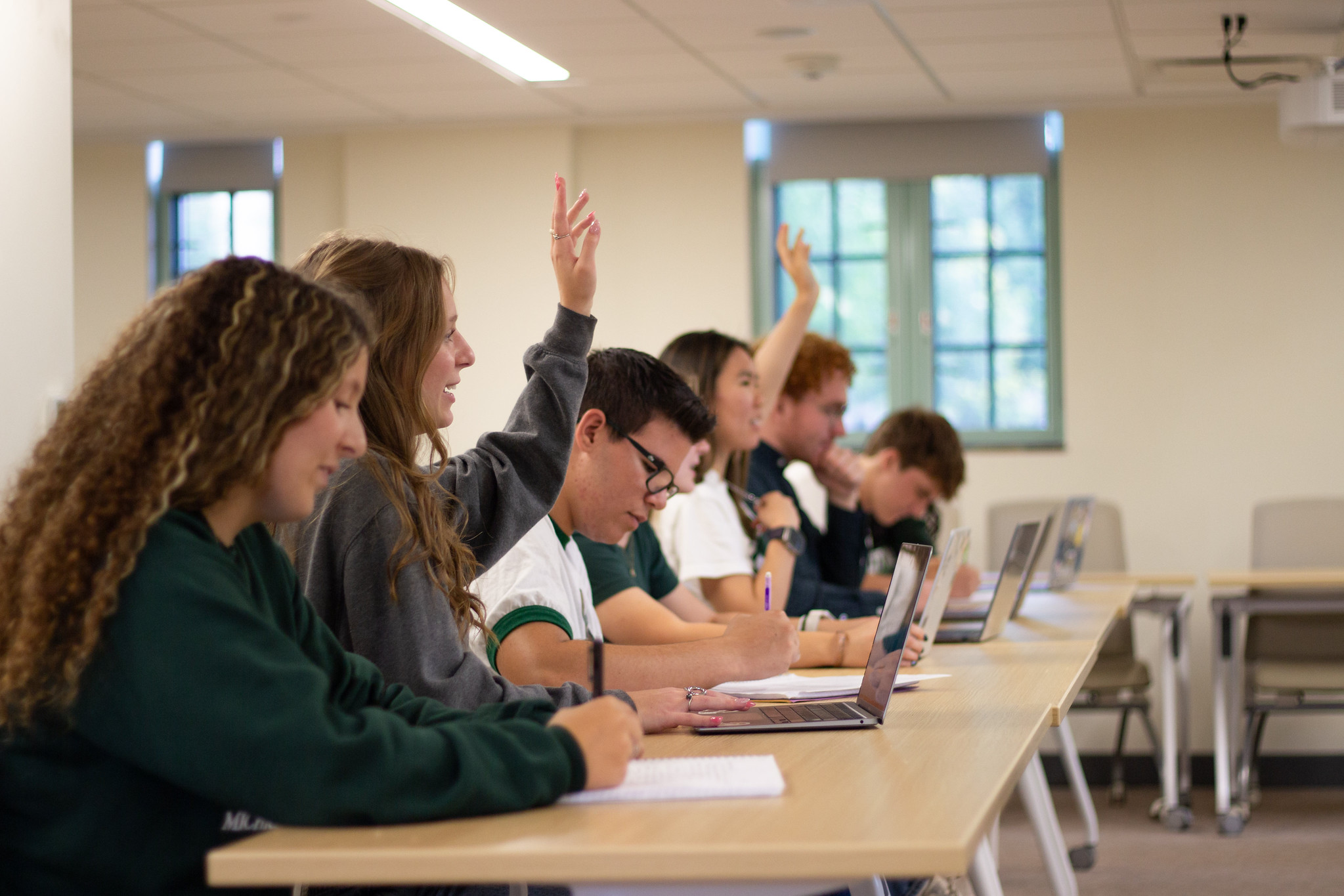 A group of Honors College students sitting in the front row of a class in the Kennicott Classroom at Campbell Hall. Two students are raising their hands, while others look intently at their notebooks and laptops.
