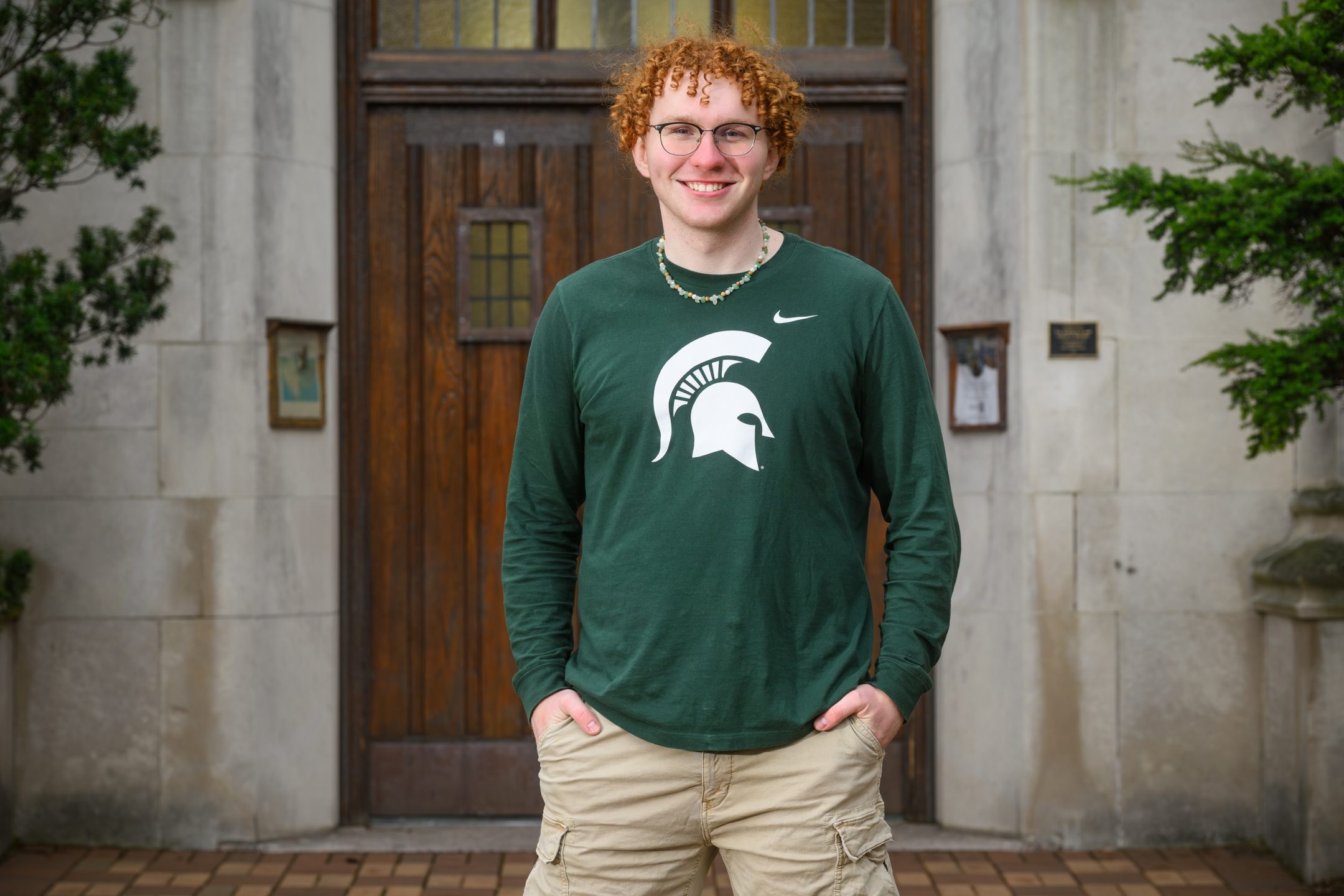 Honors College student Jagger Wraalstad standing and smiling in front of Beaumont Tower with hands in pockets, wearing a long-sleeved green and white Michigan State shirt.
