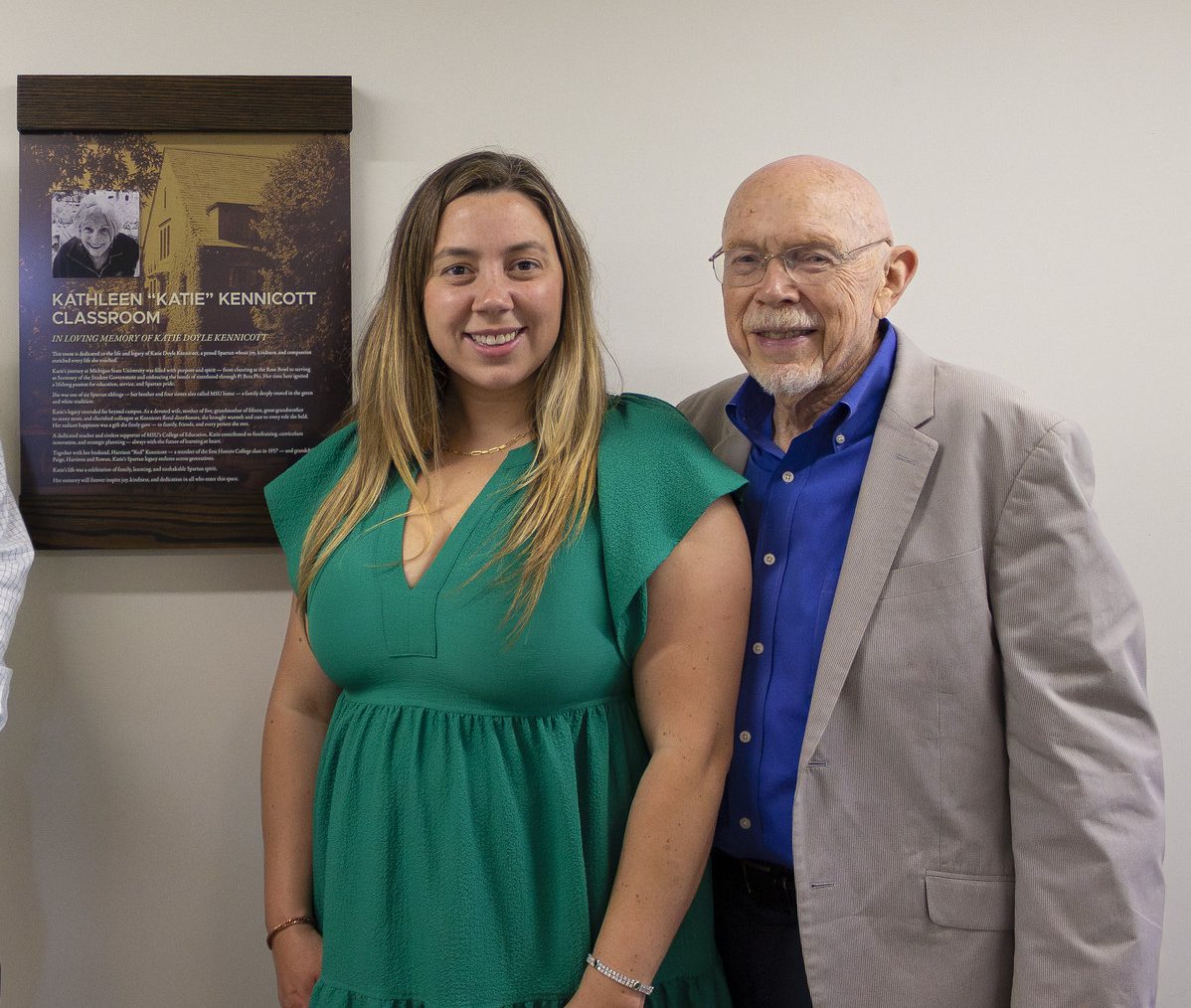 MSU Honors College alumni Paige Kennicott Jacques and Red Kennicott in dressy attire, standing next to the plaque for the Kathleen "Katie" Kennicott Classroom in MSU's Campbell Hall.
