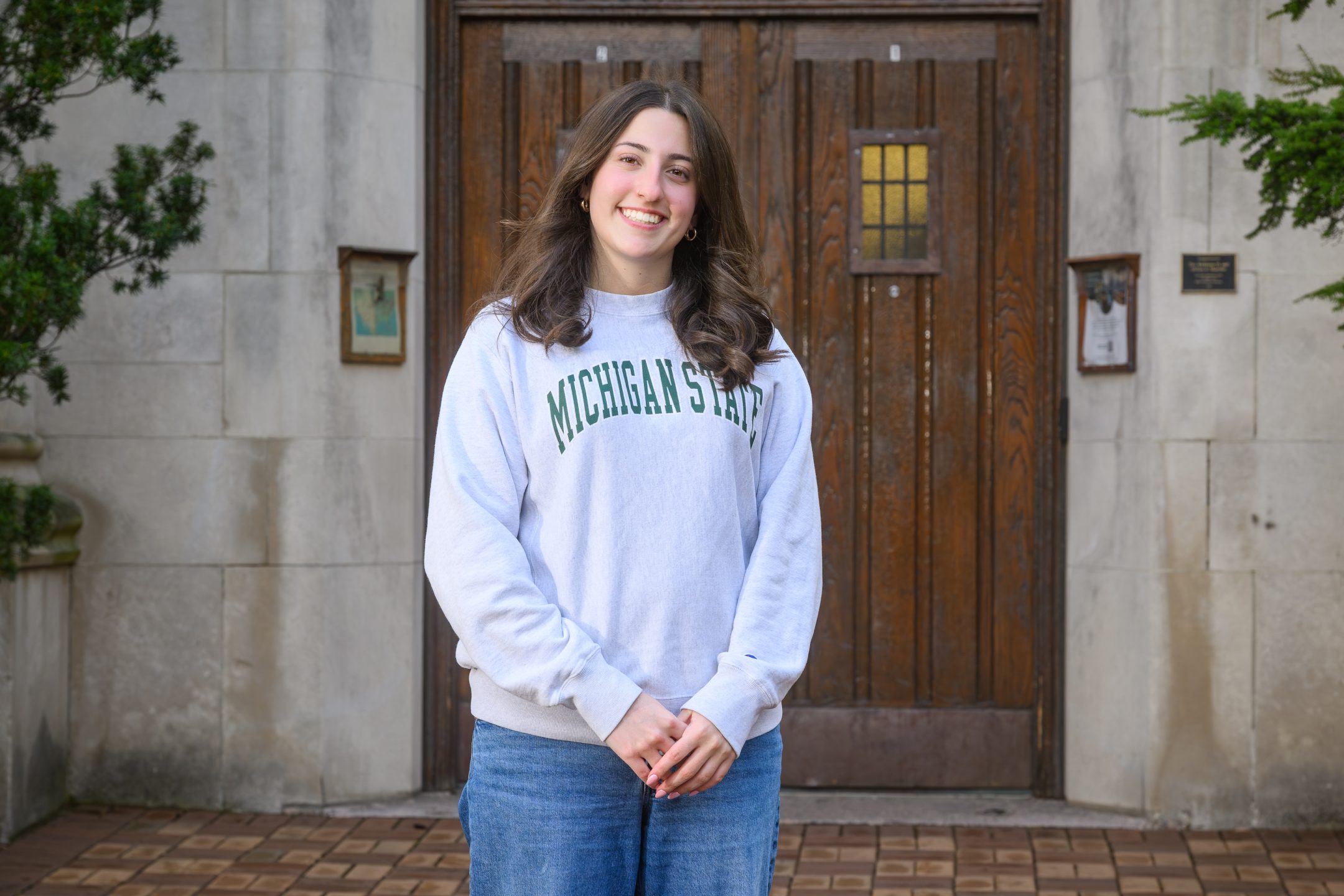 Honors College student Maddie Hanes standing and smiling in front of Beaumont Tower with hands in front of pockets, wearing a green and grey Michigan State sweater.