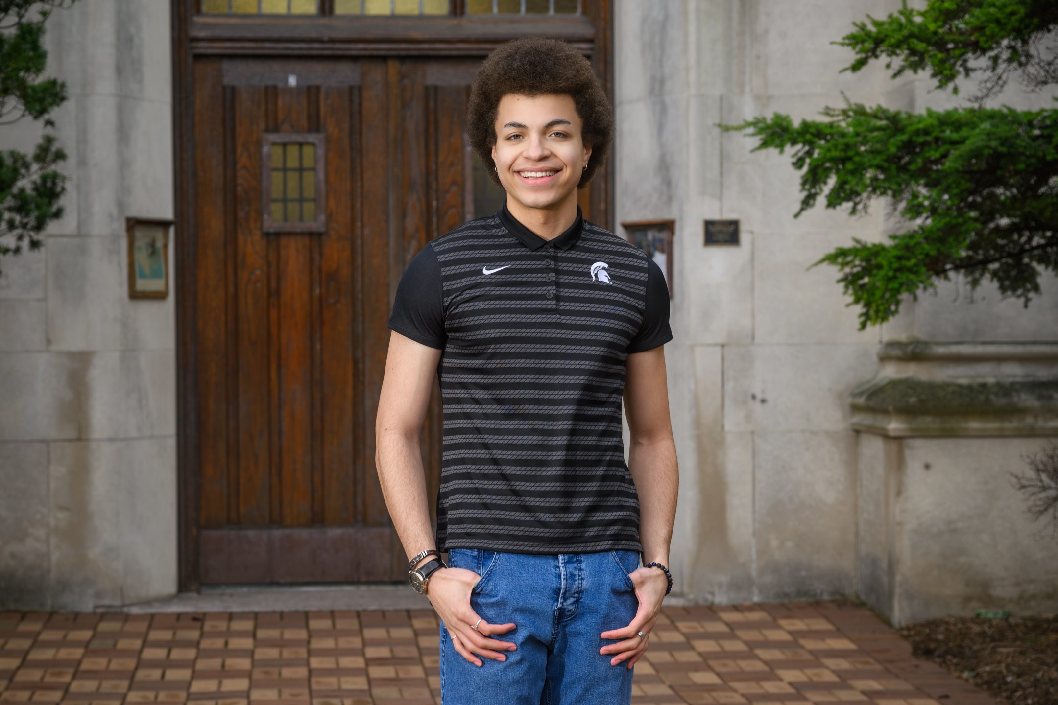 Honors College student Roberto Marcos Garcia standing and smiling in front of Beaumont Tower with hands in pockets, wearing a gray and black Michigan State polo.