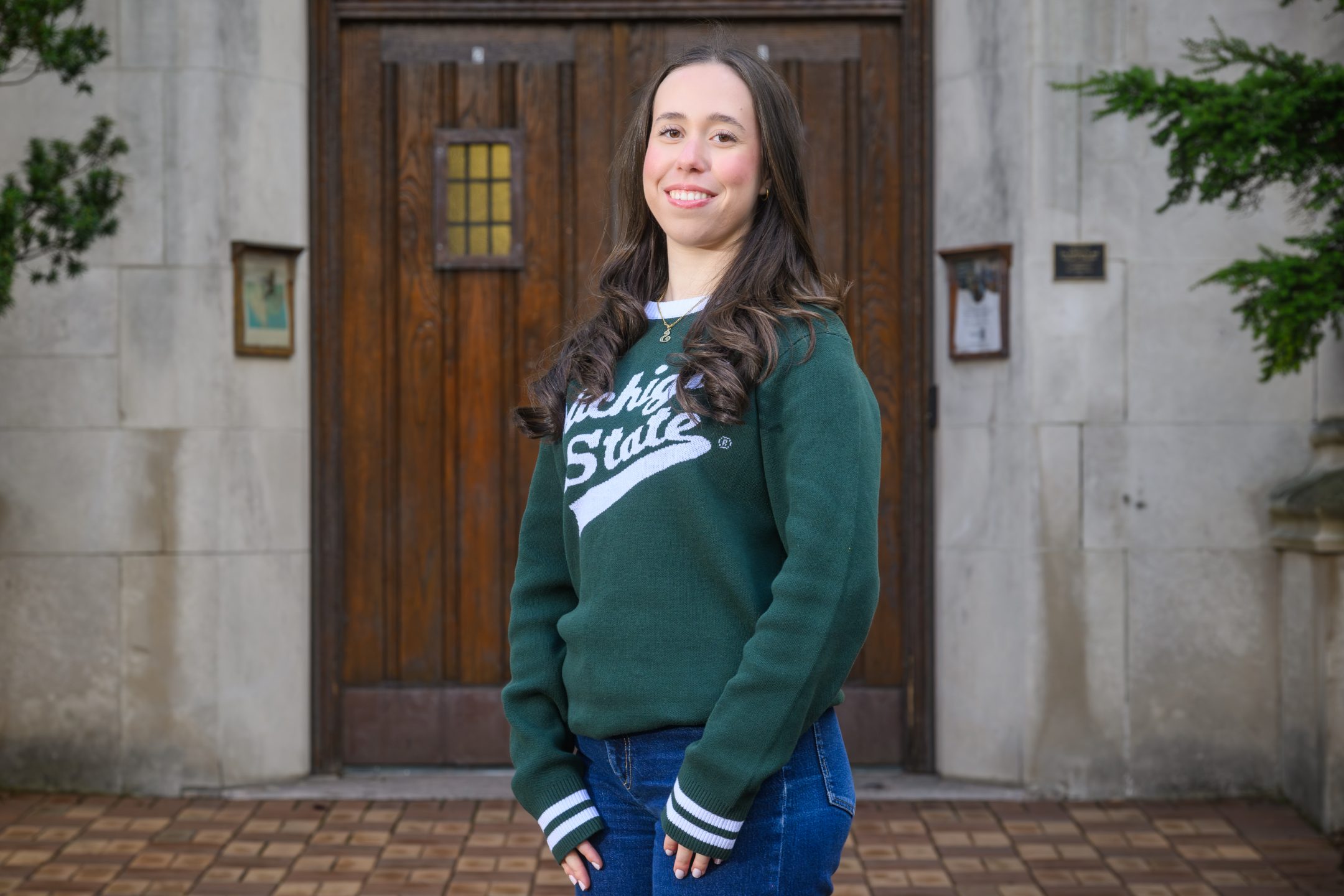 Honors College student Sarah Bejleri standing and smiling in front of Beaumont Tower with hands down, wearing a green and white Michigan State sweater.