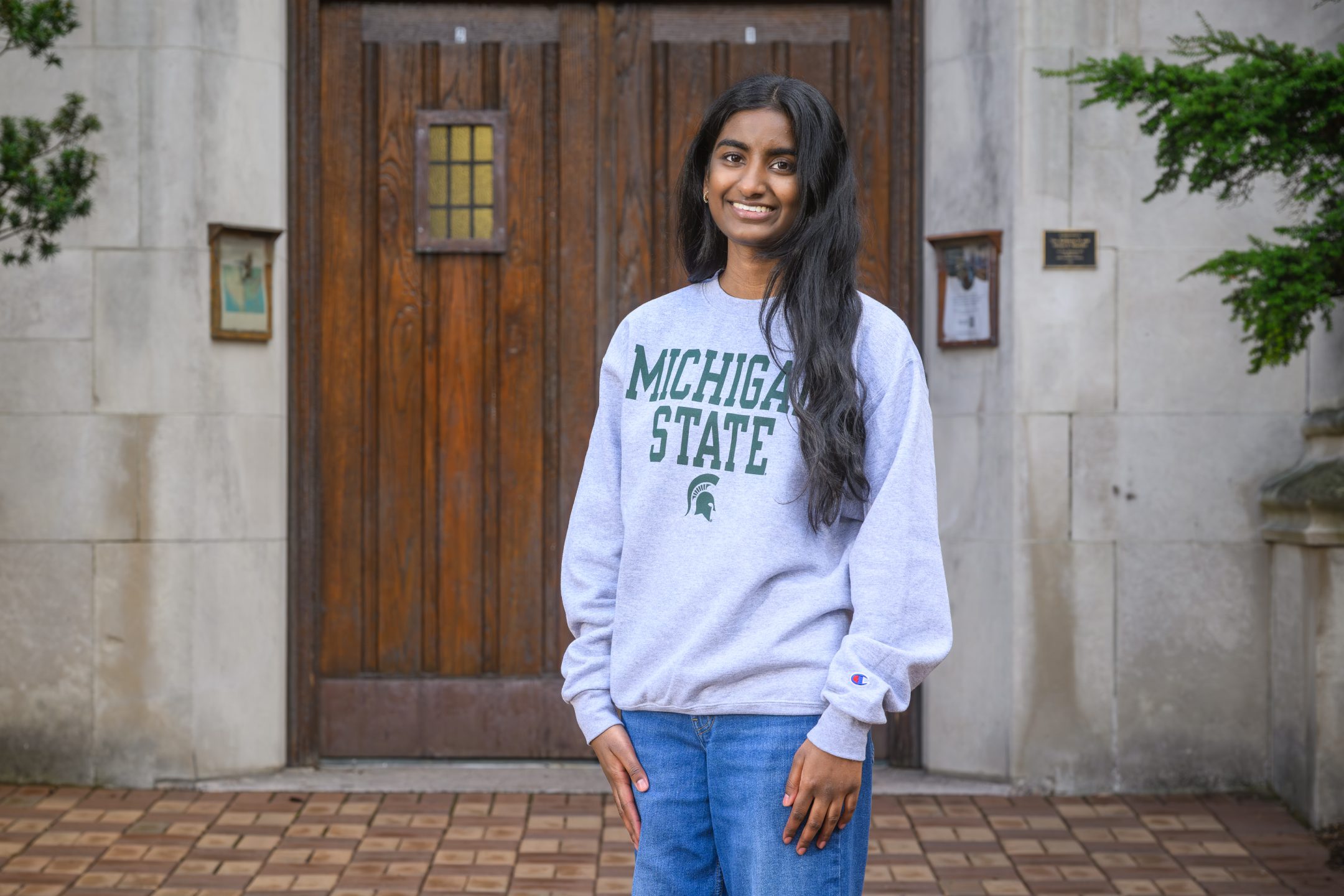 Honors College student Shrinidhi Pola standing and smiling in front of Beaumont Tower with hands in front of pockets, wearing a green and grey Michigan State sweater.