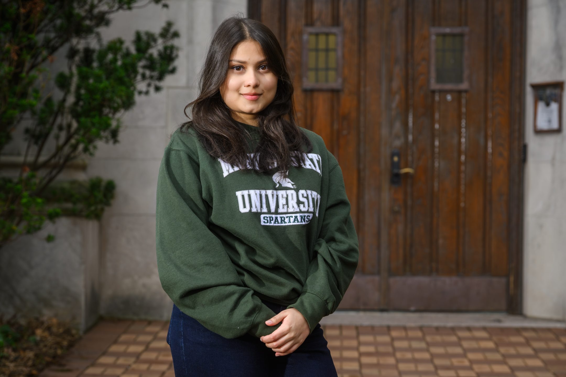 Honors College student Sumaiya Imad standing and softly smiling in front of Beaumont Tower with hands folded, wearing a green and white Michigan State sweater.