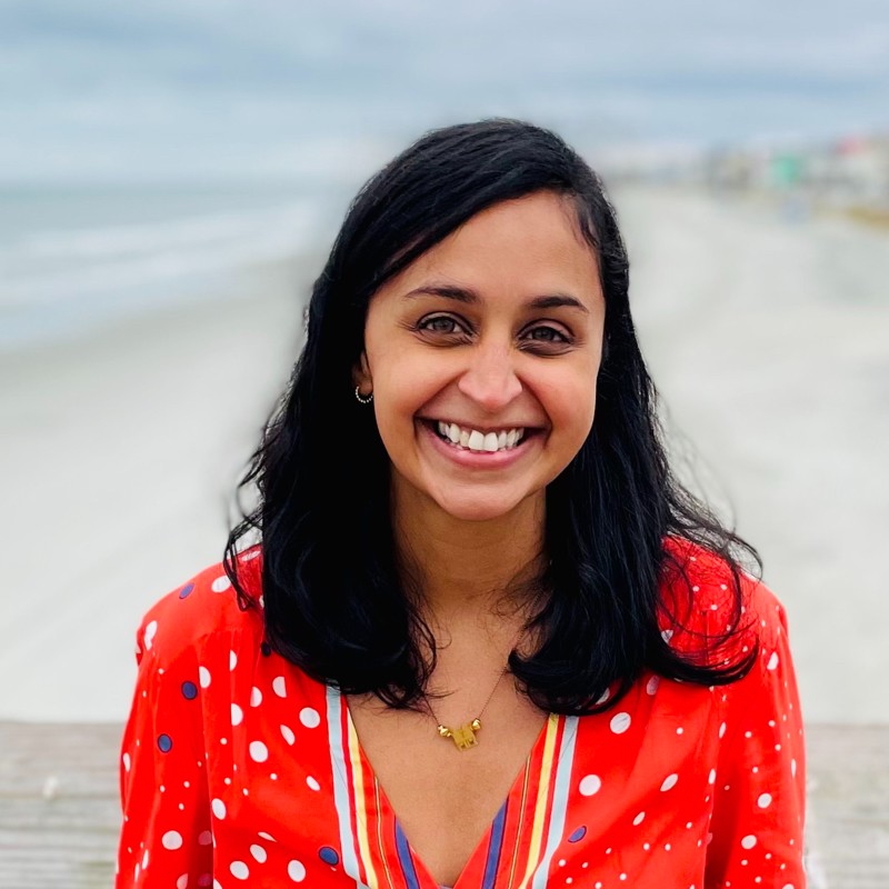 Michigan State University Honors College alumna Varsha Monick smiling in a bright red patterned shirt. A blurred body of water is in the background.